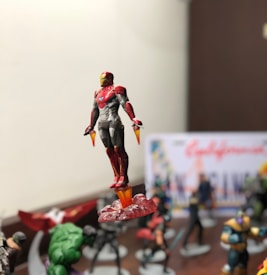 A detailed figure of a superhero in red and gold armor is prominently displayed, seemingly in flight with flames appearing beneath its feet. The background is filled with other superhero figures, slightly blurred, suggesting a collection or display setting.