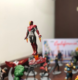 A detailed figure of a superhero in red and gold armor is prominently displayed, seemingly in flight with flames appearing beneath its feet. The background is filled with other superhero figures, slightly blurred, suggesting a collection or display setting.