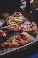 Slices of pepperoni pizza are served in a cardboard box alongside a portion of French fries. Small containers of dipping sauces, including one with a tomato-based sauce and another likely containing a mustard or cheese sauce, are visible. The pizza crust is slightly browned and crispy, with melted cheese and pepperoni slices on top. The scene is cozy and suggests a casual dining experience.