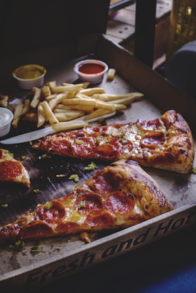 Slices of pepperoni pizza are served in a cardboard box alongside a portion of French fries. Small containers of dipping sauces, including one with a tomato-based sauce and another likely containing a mustard or cheese sauce, are visible. The pizza crust is slightly browned and crispy, with melted cheese and pepperoni slices on top. The scene is cozy and suggests a casual dining experience.