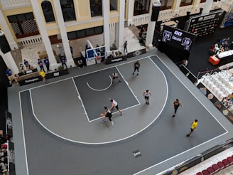 An indoor basketball game is being played on a half court surrounded by spectators. The scoreboard shows a score of 14 to 19 with 2 minutes and 20 seconds remaining. Players wearing black and white jerseys are actively engaged in the game, while referees and other officials are present. The setting appears modern with large columns and an industrial ceiling.