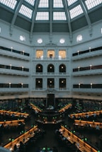 A large, domed reading room with multiple rows of desks arranged in a circular fashion around a central point. The space is filled with people studying under green reading lamps. The high ceiling features large windows, allowing natural light to filter into the room, illuminating the white walls adorned with decorative architectural elements. Shelves filled with books line the walls.