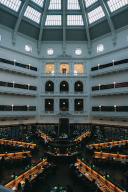 A large, domed reading room with multiple rows of desks arranged in a circular fashion around a central point. The space is filled with people studying under green reading lamps. The high ceiling features large windows, allowing natural light to filter into the room, illuminating the white walls adorned with decorative architectural elements. Shelves filled with books line the walls.