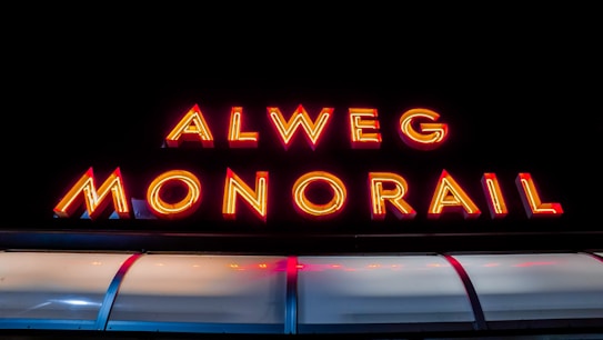 Bright neon sign displaying the words 'Alweg Monorail' in vivid red and yellow lights against a dark background. Beneath the sign, there is an illuminated curved structure that resembles the top part of a monorail train.