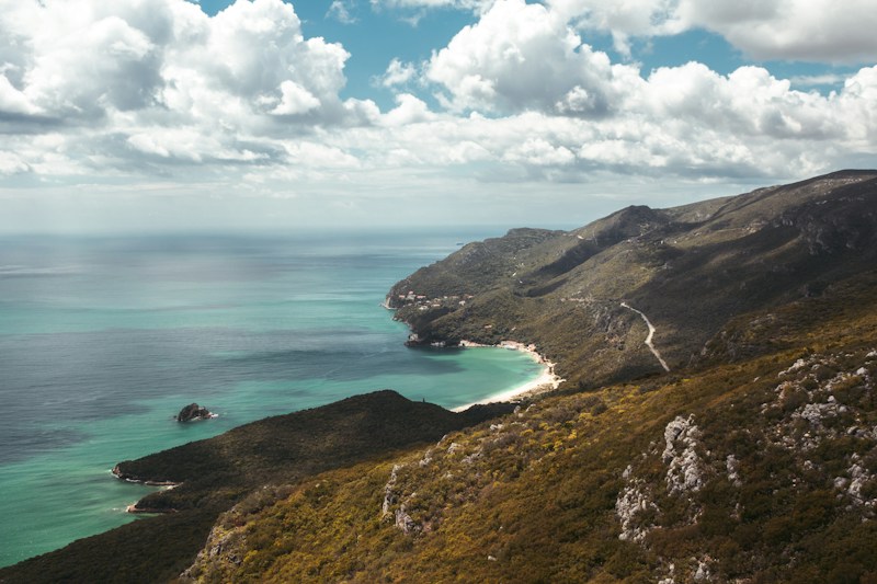 A serene coastal landscape featuring a turquoise sea bordered by a rugged coastline. Rolling hills covered with dense vegetation gently slope towards the water. A narrow, winding road traverses the hills, leading to a small, sandy beach. The sky is partly cloudy, adding a dynamic element to the tranquil scene.
