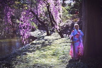 A person dressed in a kimono with floral patterns stands beside a large tree. The scene is set in a garden featuring a mix of green foliage and hanging branches of pink flowers, with a small body of water visible in the background.