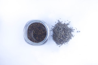 A glass jar filled with dried lavender buds is positioned next to a small pile of loose lavender on a white surface.