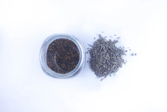 A close-up of a handmade skincare jar surrounded by lavender seeds and delicate flower petals on a wooden table.