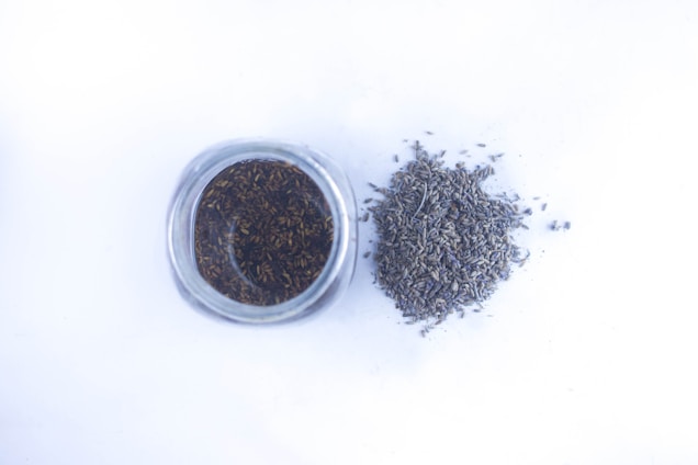 A glass jar filled with dried lavender buds is positioned next to a small pile of loose lavender on a white surface.