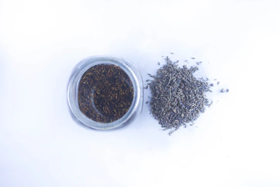 A close-up of a handmade skincare jar surrounded by lavender seeds and delicate flower petals on a wooden table.