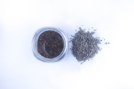 A glass jar filled with dried lavender buds is positioned next to a small pile of loose lavender on a white surface.
