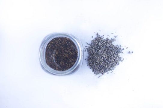 A glass jar filled with dried lavender buds is positioned next to a small pile of loose lavender on a white surface.