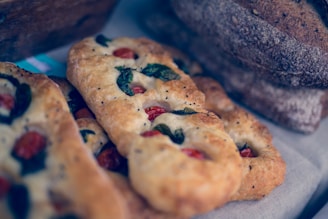 A close-up view of freshly baked flatbreads adorned with cherry tomatoes and fresh basil leaves, juxtaposed with several loaves of rustic brown bread. The texture of the bread is highlighted, showcasing a golden, crispy crust.