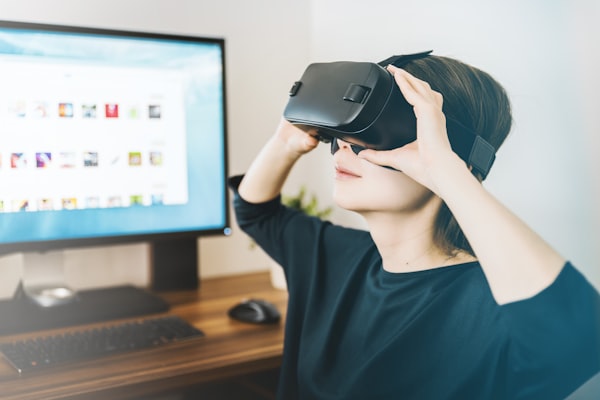 A woman wears a virtual reality headset, sitting in front of a computer screen