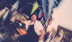 A person wearing an apron and hat is standing in front of a food stall, preparing food with items like hotdogs visible. The scene appears blurred with dynamic motion and colorful banners hang overhead, creating a festive atmosphere.