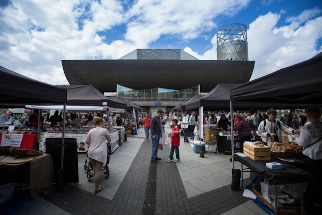A busy outdoor market scene with various vendors under black tents. Many people are walking around, browsing, and shopping. The background features a modern building with a unique architectural design, including a large angular overhang and a tower-like structure. The sky is partly cloudy with patches of blue.