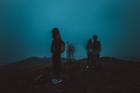 A group of people stand on a dark hill under a teal-tinted sky, with some figures appearing blurred or translucent. The setting suggests an eerie or mysterious atmosphere.