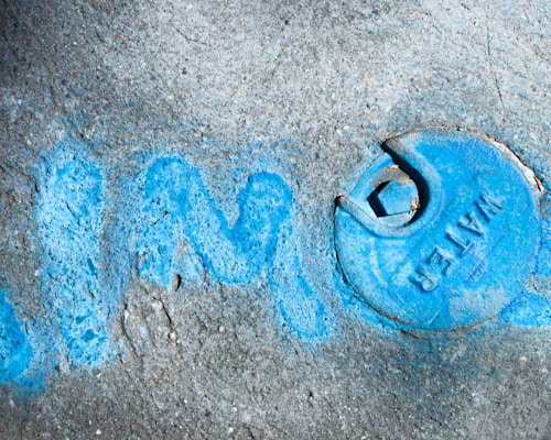 A concrete surface with a blue-colored water valve cover is partially surrounded by faint blue graffiti. The word 'WATER' is embossed on the circular cover, which is weathered with marks and scratches.