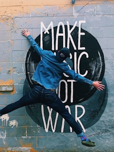 A person in denim clothing and a cap is leaping in front of a mural painted on a brick wall. The mural contains the words 'MAKE MUSIC NOT WAR' in large, white letters set against a circular, dark background. The wall shows signs of wear, with patches of yellow and gray paint.