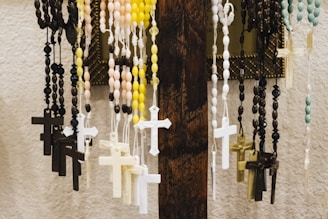 Several rosaries with beads and crucifixes hang from a wooden stand. The rosaries are various colors, including black, yellow, pink, blue, and white. The backdrop features a textured white wall and a framed item partially visible.