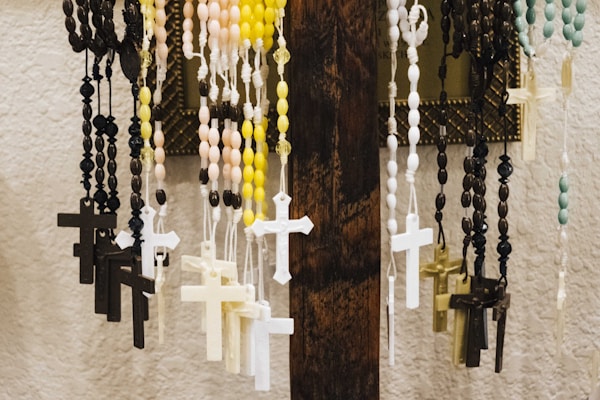 Several rosaries with beads and crucifixes hang from a wooden stand. The rosaries are various colors, including black, yellow, pink, blue, and white. The backdrop features a textured white wall and a framed item partially visible.
