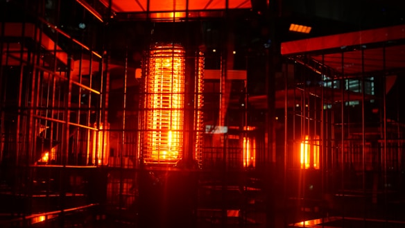 A futuristic and industrial setting with multiple glowing red-orange heat lamps enclosed in metal cages. The scene is dark, with a strong contrast between the vivid light sources and their metallic surroundings, creating a warm, almost intense glow.
