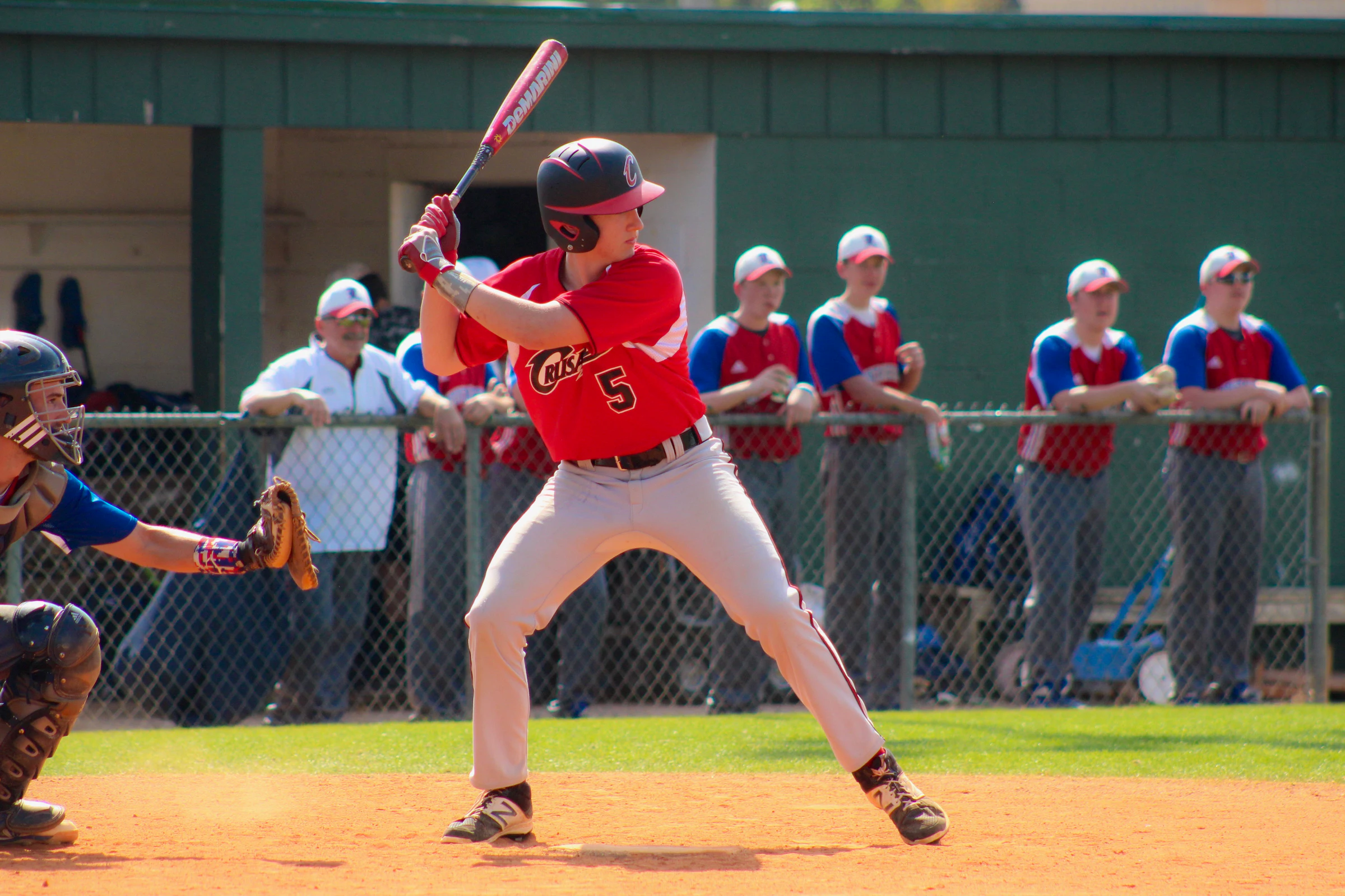 A hitter stands in at the plate while teammates lean on the fence along the dugout watching the at-bat.