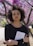 A person with curly hair stands outdoors holding a book titled '101 Essays that will Change the way You Think.' They are wearing a long-sleeve black top and stand amidst trees with vibrant pink flowers on a sunny day.