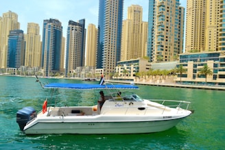A small yacht navigates a tranquil marina with a skyline of tall, modern skyscrapers in the background. The boat features a blue canopy and is adorned with a flag.