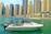 A small yacht navigates a tranquil marina with a skyline of tall, modern skyscrapers in the background. The boat features a blue canopy and is adorned with a flag.