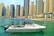 A small yacht navigates a tranquil marina with a skyline of tall, modern skyscrapers in the background. The boat features a blue canopy and is adorned with a flag.