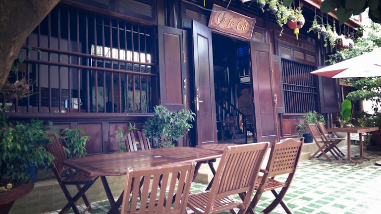 A cozy outdoor cafe setting with wooden furniture, green plants, and traditional architecture. The entrance features open doors and a sign above. Wooden chairs and tables are neatly arranged on a tiled floor with some potted plants nearby. An umbrella provides shade in the corner.
