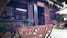 A cozy outdoor cafe setting with wooden furniture, green plants, and traditional architecture. The entrance features open doors and a sign above. Wooden chairs and tables are neatly arranged on a tiled floor with some potted plants nearby. An umbrella provides shade in the corner.