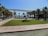 A landscaped area with a central green lawn surrounded by palm trees and paved walkways. There is a modern architectural structure providing shade, and in the background, a view of the waterfront with several people and buildings visible.