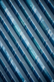 Diagonal rows of glossy, blue-tinted glass panels create a geometric pattern with reflective surfaces. The panels capture light and shadows, adding depth and complexity to the design.