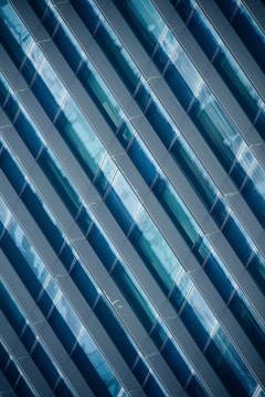 Diagonal rows of glossy, blue-tinted glass panels create a geometric pattern with reflective surfaces. The panels capture light and shadows, adding depth and complexity to the design.