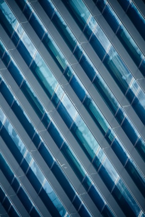 Diagonal rows of glossy, blue-tinted glass panels create a geometric pattern with reflective surfaces. The panels capture light and shadows, adding depth and complexity to the design.