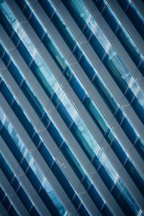 Diagonal rows of glossy, blue-tinted glass panels create a geometric pattern with reflective surfaces. The panels capture light and shadows, adding depth and complexity to the design.