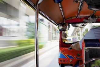 An interior view of a tuk-tuk with a driver visible in the rearview mirror. The vehicle is in motion, with blurred buildings and greenery outside, indicating speed. The interior elements of the tuk-tuk, such as the roof, poles, and part of the driver's seat, are visible.