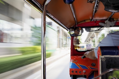 An interior view of a tuk-tuk with a driver visible in the rearview mirror. The vehicle is in motion, with blurred buildings and greenery outside, indicating speed. The interior elements of the tuk-tuk, such as the roof, poles, and part of the driver's seat, are visible.