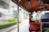 An interior view of a tuk-tuk with a driver visible in the rearview mirror. The vehicle is in motion, with blurred buildings and greenery outside, indicating speed. The interior elements of the tuk-tuk, such as the roof, poles, and part of the driver's seat, are visible.