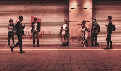 Several young adults are standing against a shuttered storefront in an urban setting, each engaged with their mobile devices or interacting with one another. The setting appears to be a city street during the evening, as indicated by the warm artificial lighting.
