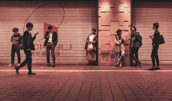 Several young adults are standing against a shuttered storefront in an urban setting, each engaged with their mobile devices or interacting with one another. The setting appears to be a city street during the evening, as indicated by the warm artificial lighting.