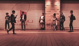 Several young adults are standing against a shuttered storefront in an urban setting, each engaged with their mobile devices or interacting with one another. The setting appears to be a city street during the evening, as indicated by the warm artificial lighting.