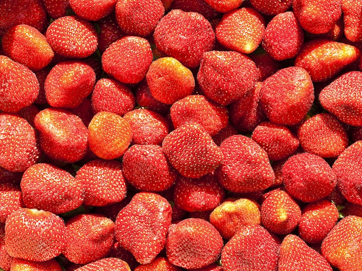 A densely packed collection of fresh, ripe strawberries with vibrant red skin and visible seeds. The strawberries are evenly distributed across the image, showcasing their varying sizes and shapes. The surface glistens slightly, suggesting freshness.
