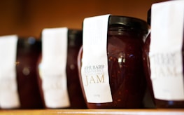 Several jars of rhubarb raspberry vanilla bean jam lined up on a shelf, each adorned with a white label. The jars are tightly sealed with black lids, and the contents appear to be thick and rich in texture, with visible seeds suggesting a homemade quality.