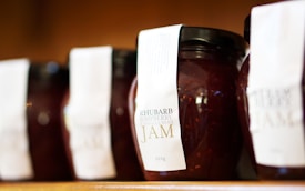 Several jars of rhubarb raspberry vanilla bean jam lined up on a shelf, each adorned with a white label. The jars are tightly sealed with black lids, and the contents appear to be thick and rich in texture, with visible seeds suggesting a homemade quality.