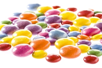 A collection of brightly colored, glossy candies scattered on a white background. The candies vary in size and exhibit a range of colors, including shades of yellow, orange, red, purple, blue, and green. They have a smooth, shiny surface that reflects light, giving them an appealing and vibrant look.