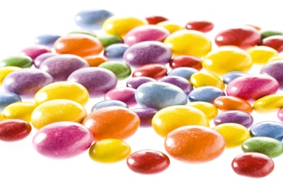 A collection of brightly colored, glossy candies scattered on a white background. The candies vary in size and exhibit a range of colors, including shades of yellow, orange, red, purple, blue, and green. They have a smooth, shiny surface that reflects light, giving them an appealing and vibrant look.