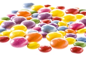 A collection of brightly colored, glossy candies scattered on a white background. The candies vary in size and exhibit a range of colors, including shades of yellow, orange, red, purple, blue, and green. They have a smooth, shiny surface that reflects light, giving them an appealing and vibrant look.